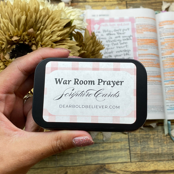 Hand holding a 'War Room Prayer' card in front of an open book on a wooden surface.