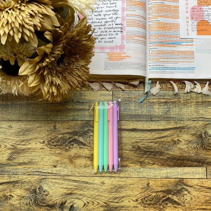 Open book with bookmarks on a wooden surface next to dried flowers and colorful pen.