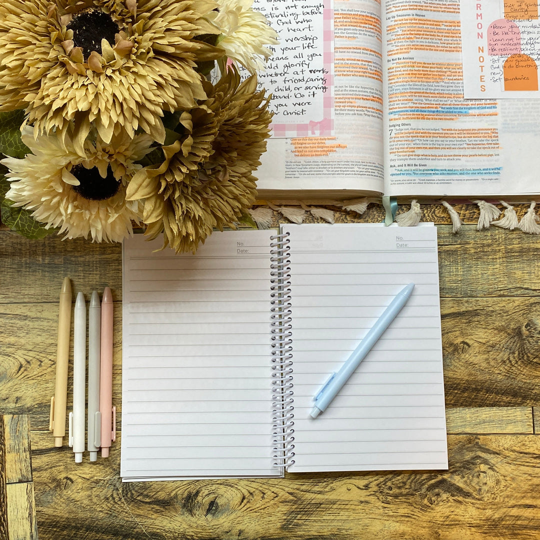 Open notebook with pen, flowers, and open book on a wooden desk