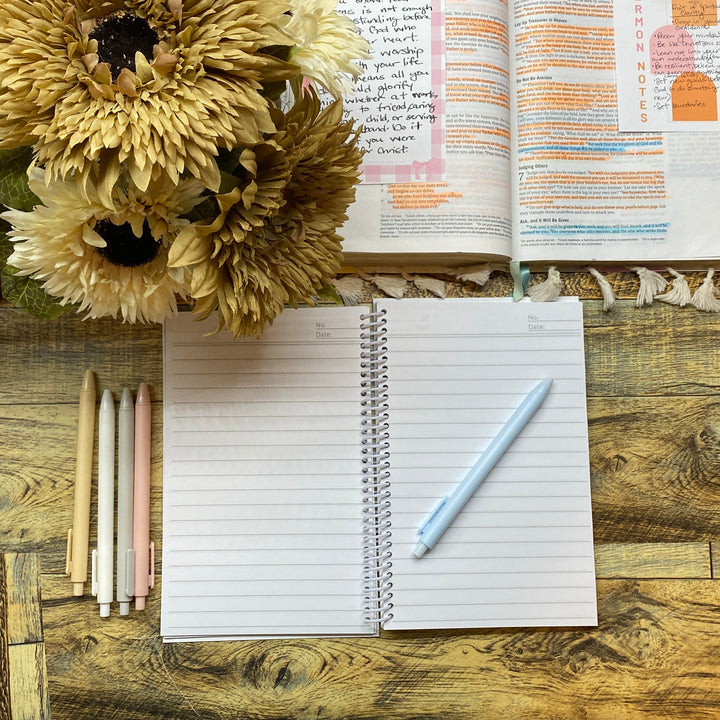 Open notebook with pen, flowers, and open book on a wooden desk