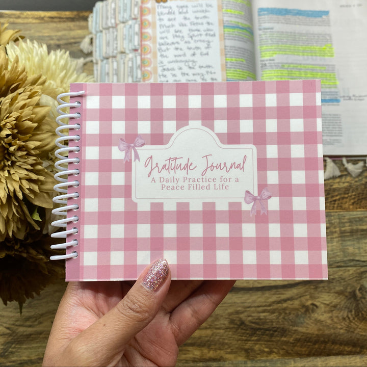 Pink checkered gratitude journal held by a hand on a wooden surface with flowers in the background