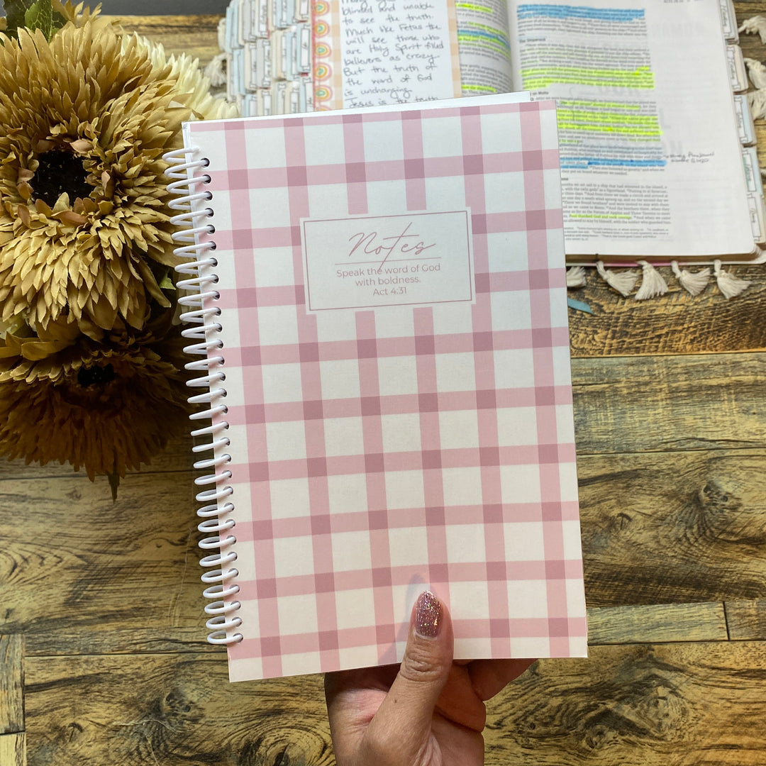 Pink checkered notebook held by a hand on a wooden surface with sunflowers and another notebook in the background.