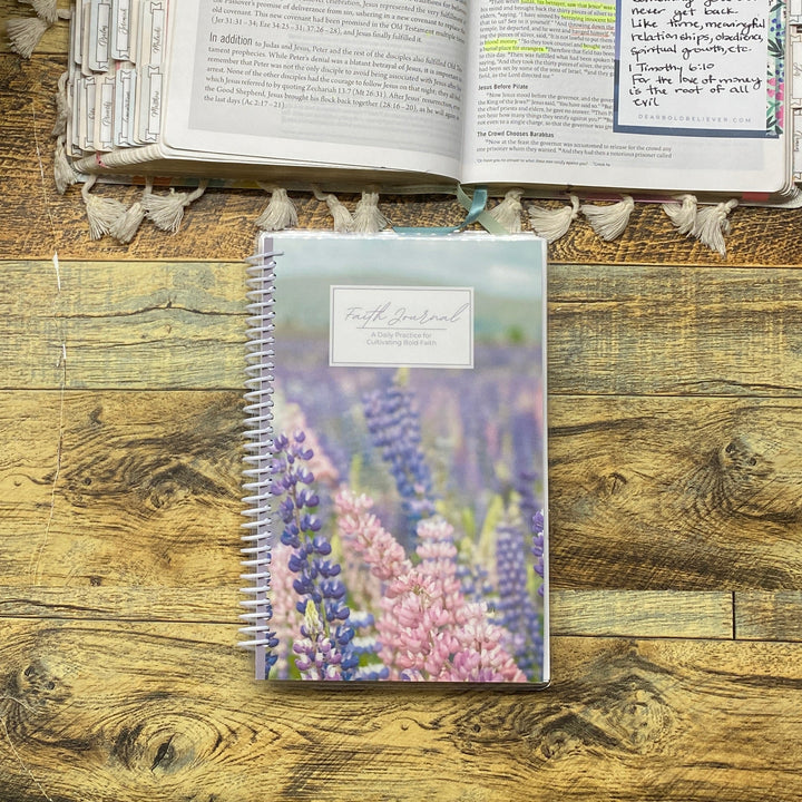 Open book with floral decorations on a wooden surface next to a spiral-bound notebook.