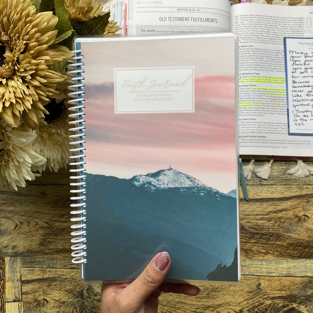Hand holding a spiral-bound notebook with a scenic cover on a wooden surface with flowers and stationery.