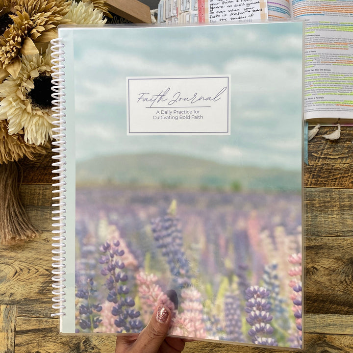 Notebook with a scenic cover held by a hand on a wooden surface with flowers and another notebook.
