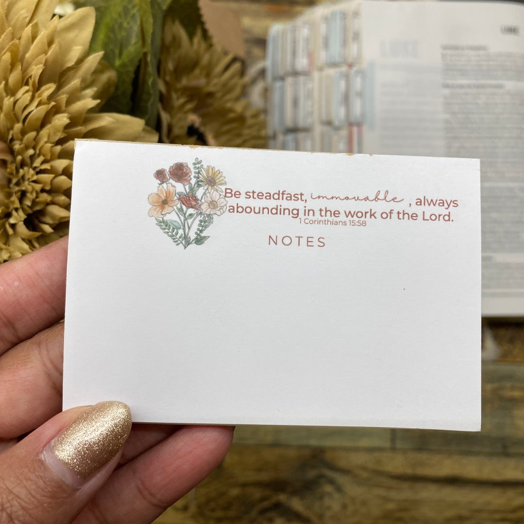 Hand holding a card with floral design and text, with sunflowers and an open book in the background.