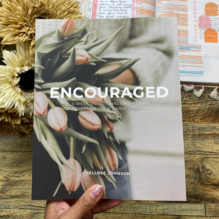 Book titled 'Encouraged' held by a hand on a wooden surface with flowers.