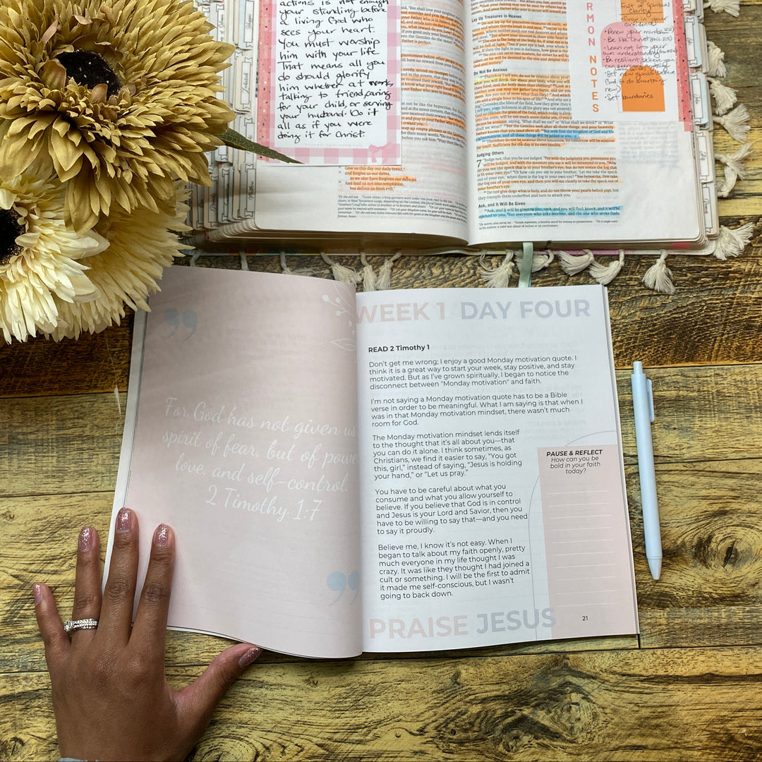 Open book with a hand on a wooden surface, next to flowers and a pen.