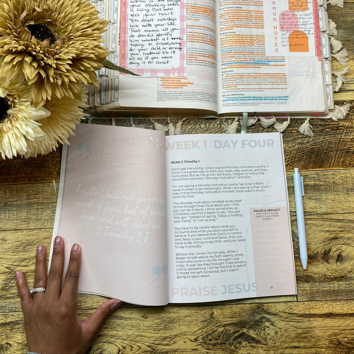 Open book with a hand on a wooden surface, next to flowers and a pen.