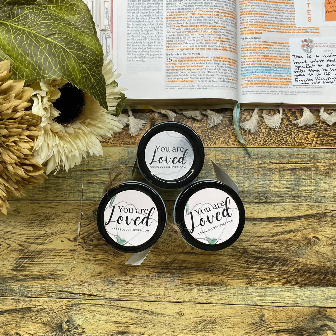 Three small black containers with 'You are Loved' text on a wooden surface with flowers and an open book.