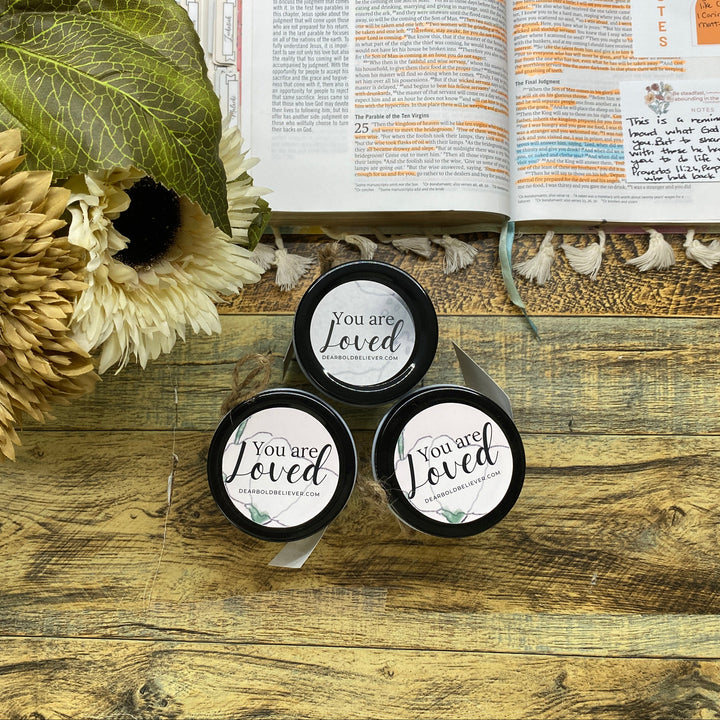 Three small black containers with 'You are Loved' text on a wooden surface with flowers and an open book.