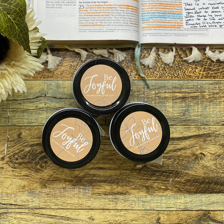 Three small black containers with 'Not Forged' text on a wooden surface with flowers and an open book in the background.
