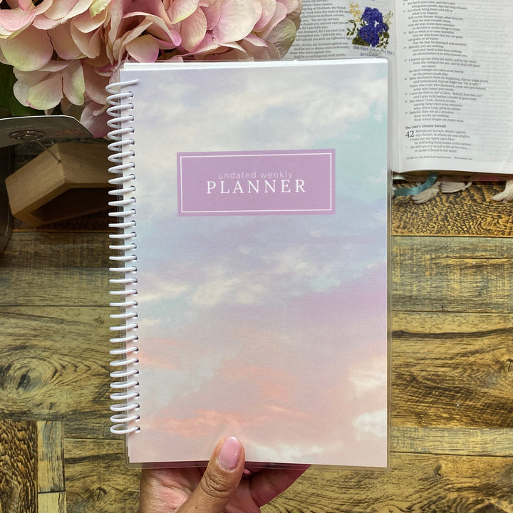 Hand holding a planner with a scenic cover on a wooden surface with flowers and books.