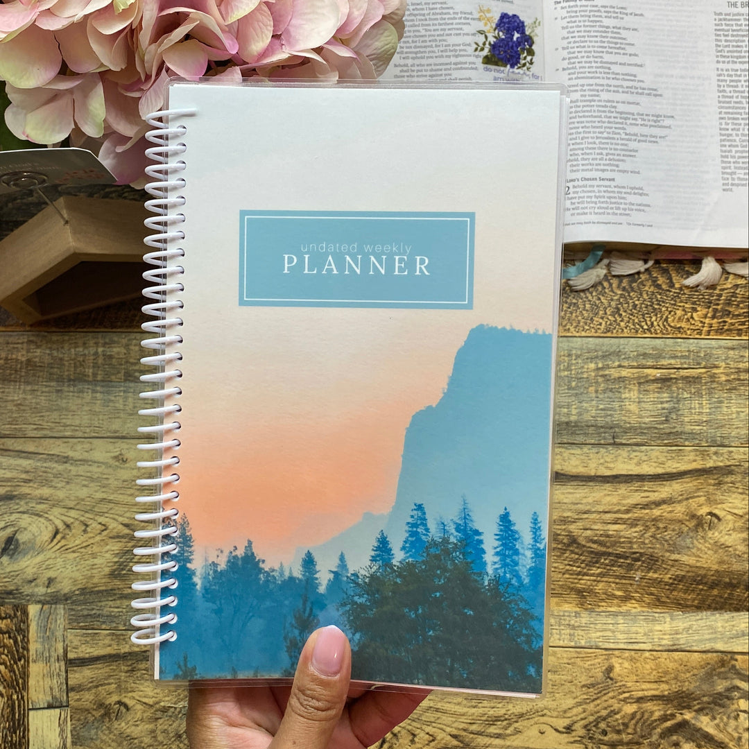 Hand holding a planner with a scenic cover on a wooden desk with flowers and a book.