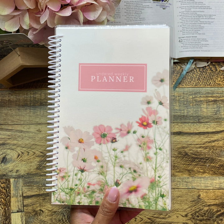 Hand holding a floral planner on a wooden desk with an open book and pink flowers.