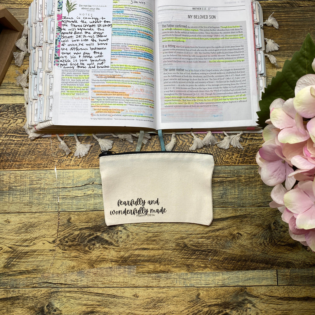 A beige fabric pouch with the words 'faithfully and wonderfully made' in black script, placed on a rustic wooden table next to an open Bible and pink hydrangea flowers.