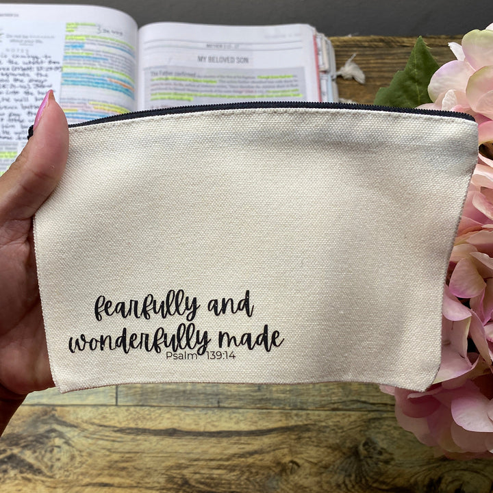 A hand holds a cream-colored fabric pouch with black script text reading 'faithfully and wonderfully made' and a smaller 'God is love' underneath, set against a rustic wooden desk background with a planner and pink flowers.
