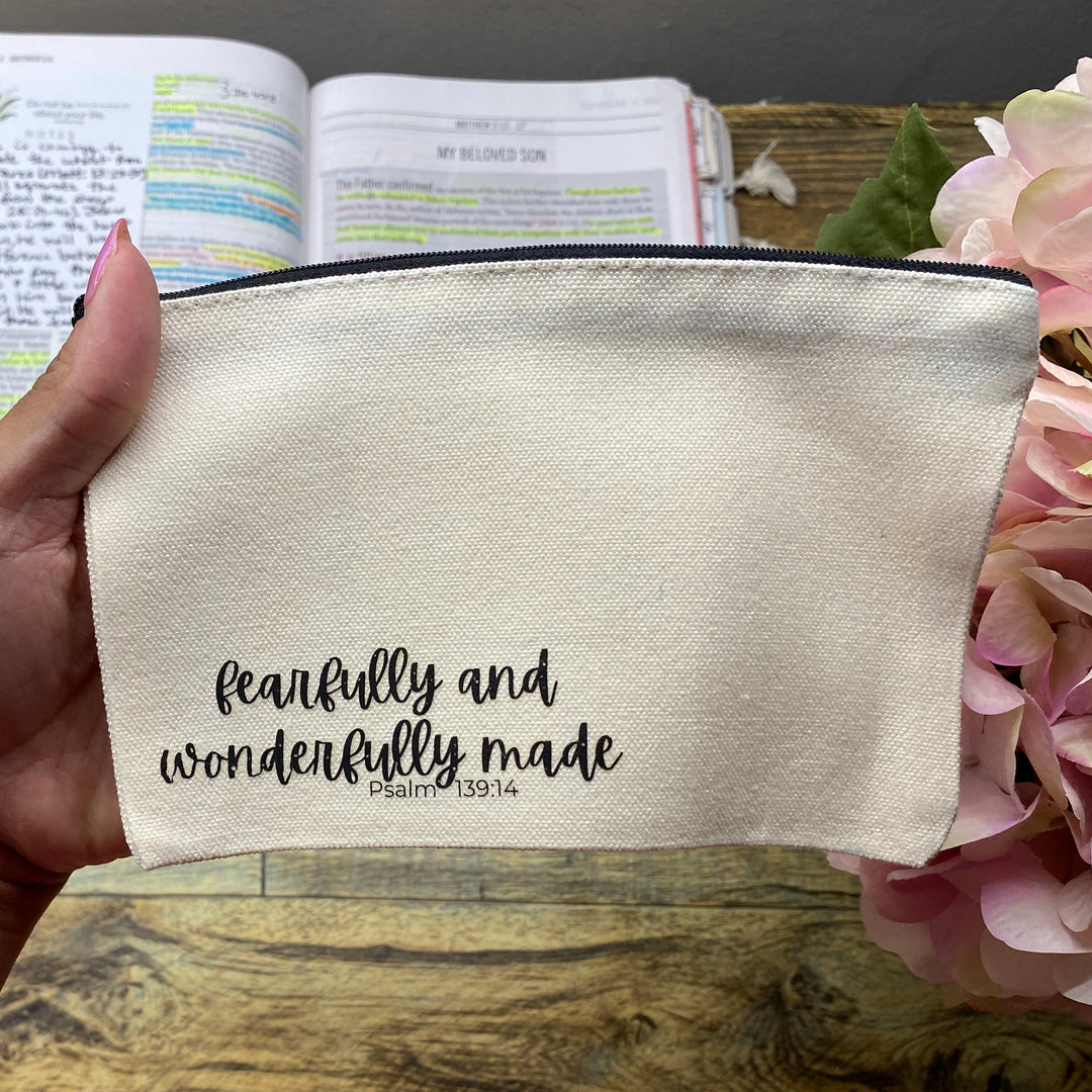 A hand holds a cream-colored fabric pouch with black script text reading 'faithfully and wonderfully made' and a smaller 'God is love' underneath, set against a rustic wooden desk background with a planner and pink flowers.
