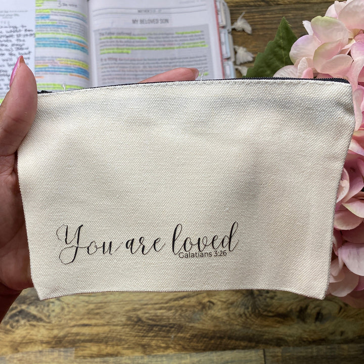 A hand holds a cream-colored fabric pouch with the words 'You are loved Galatians 6:28' printed in black script. The background shows a wooden desk with an open planner, a small cardboard box, and pink flowers.