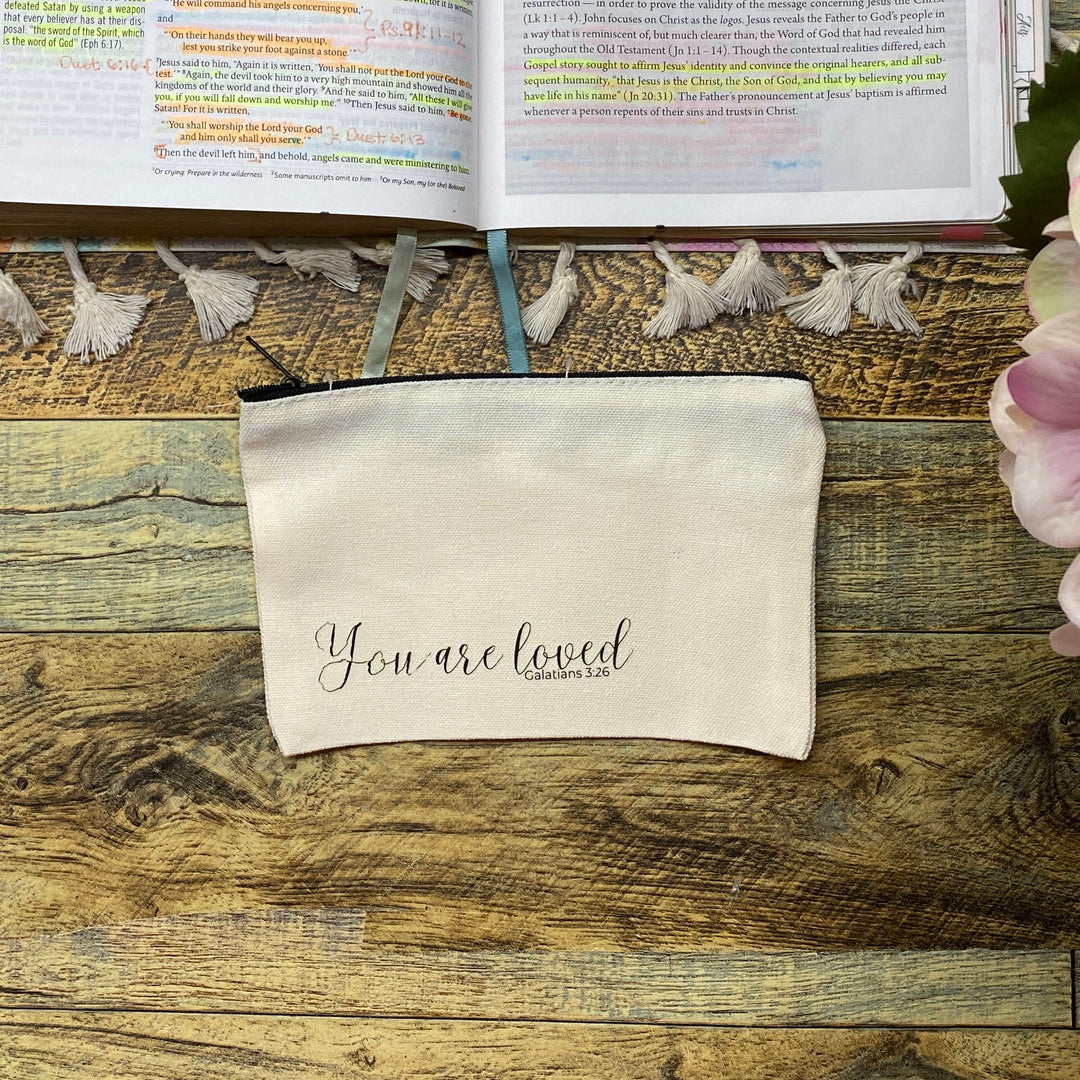 An open journal filled with colorful sticky notes and handwritten notes, displayed on a rustic wooden table alongside a cream canvas pouch that reads 'you are loved' and pink hydrangea flowers in the foreground.