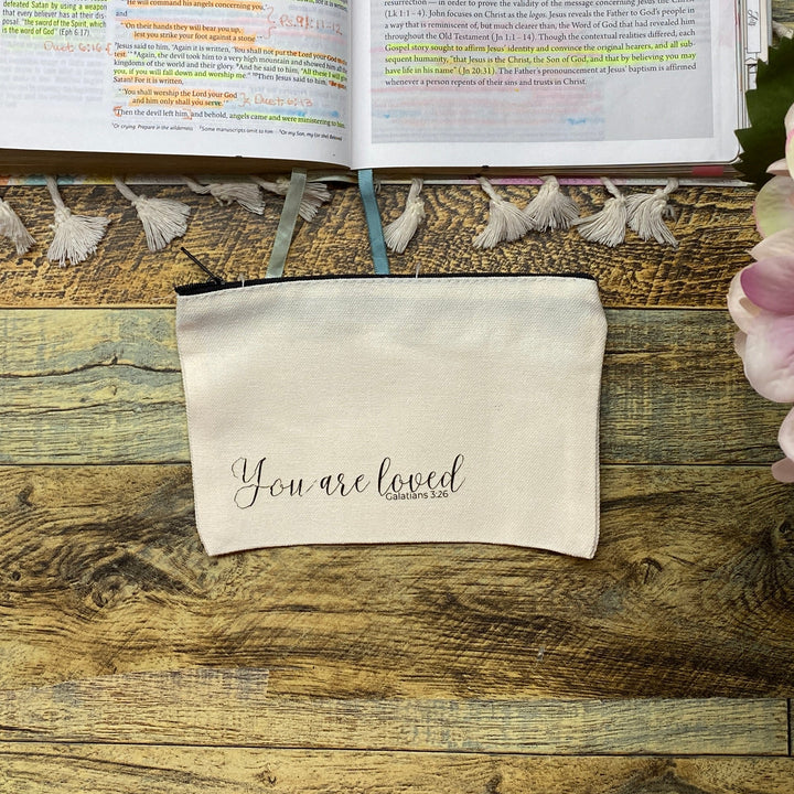 An open journal filled with colorful sticky notes and handwritten notes, displayed on a rustic wooden table alongside a cream canvas pouch that reads 'you are loved' and pink hydrangea flowers in the foreground.