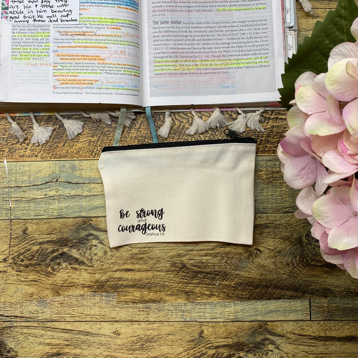 A cream canvas pouch with 'be strong campaigners' printed in black script, displayed on a rustic wooden table alongside an open journal, a pink hydrangea, and a small wooden box.