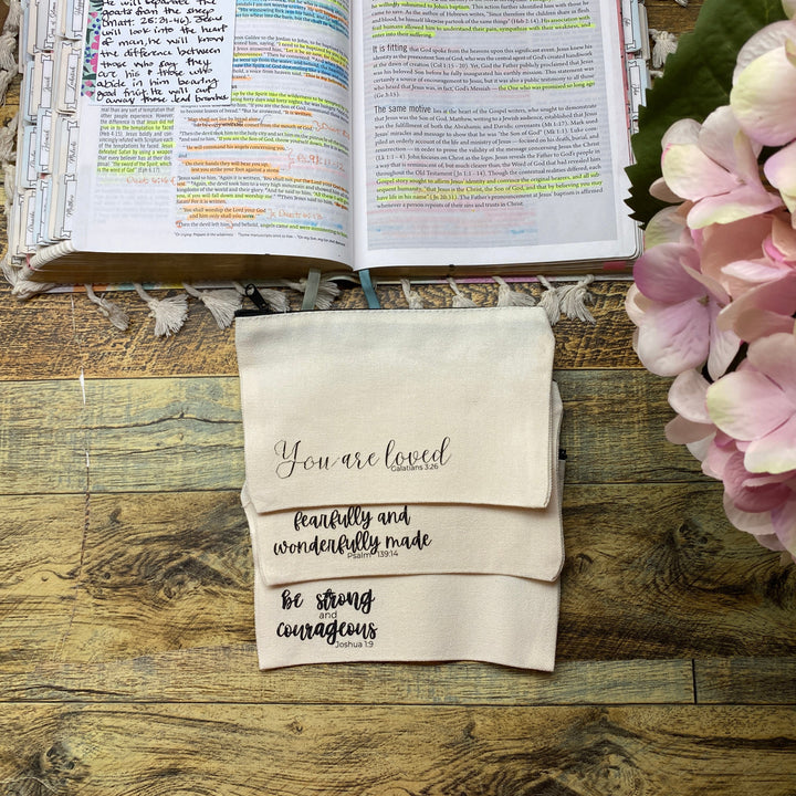 Three cream-colored fabric napkins with black cursive text are stacked on a rustic wooden table next to an open Bible and pink hydrangeas. The napkins read 'You are loved', 'beautifully and wonderfully made', and 'be strong courageous'.