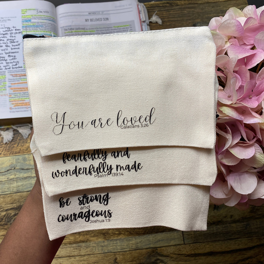 A hand holds three cream-colored fabric prayer mats stacked diagonally, printed with black inspirational Bible verses. The background shows a rustic wooden table with an open Bible, a small wooden box, and pink hydrangea flowers.