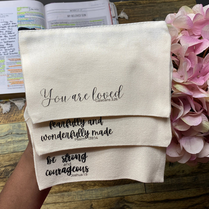 A hand holds three cream-colored fabric prayer mats stacked diagonally, printed with black inspirational Bible verses. The background shows a rustic wooden table with an open Bible, a small wooden box, and pink hydrangea flowers.
