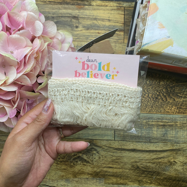 A hand holding a packaged cream-colored lace headband with a pink card that reads 'dear bold believer'. The background features a rustic wooden table, a pink hydrangea, and a notebook.