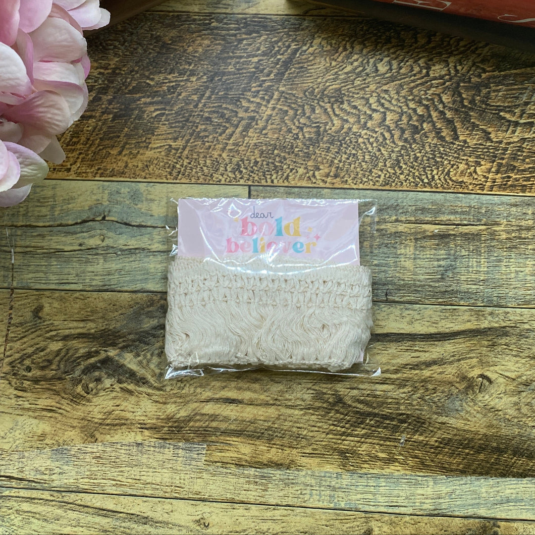 A small clear plastic bag containing a beige woven or knotted cord bundle, placed on a rustic wooden tabletop. A pink flower, a knife, and stacked papers are visible around it as props.