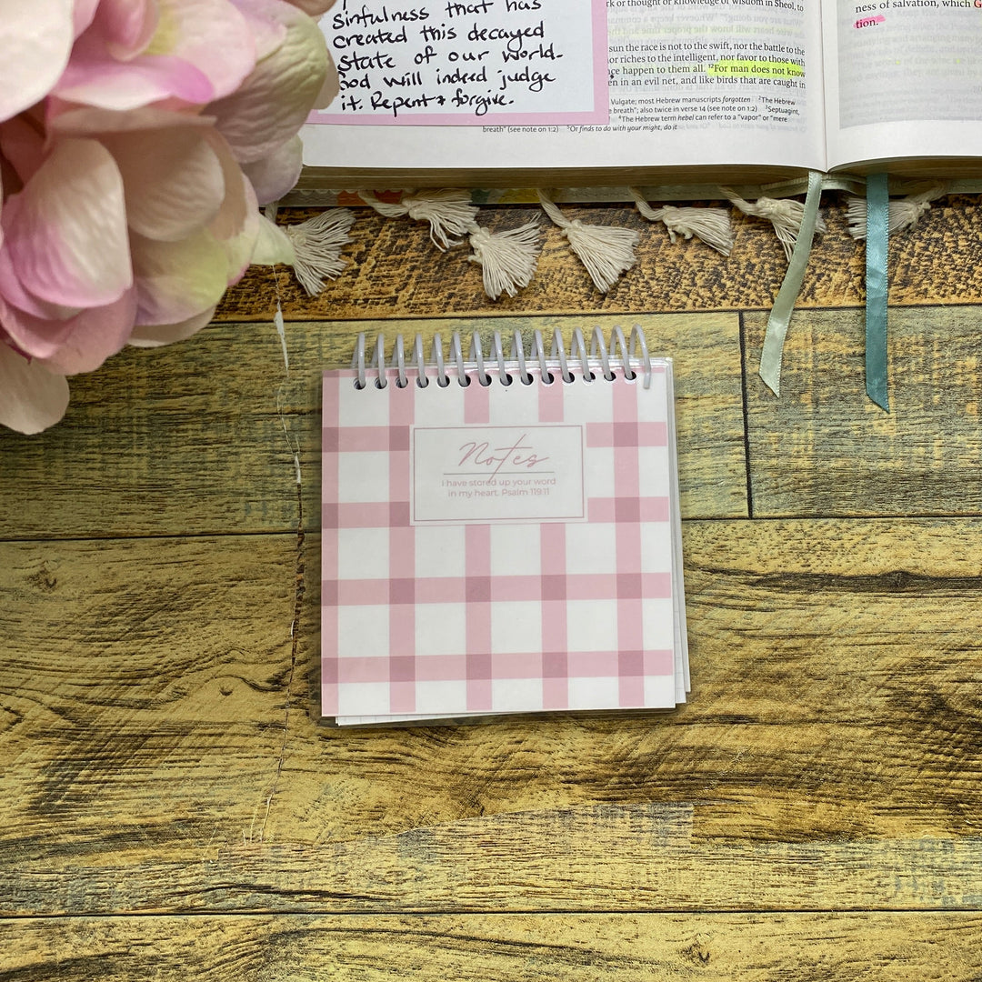 A pink and white gingham spiral notebook lies on a rustic wooden tabletop next to an open handwritten planner or journal and a bouquet of pink flowers.