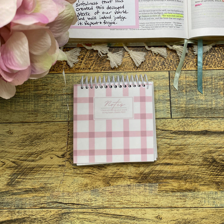 A pink and white gingham spiral notebook lies on a rustic wooden tabletop next to an open handwritten planner or journal and a bouquet of pink flowers.