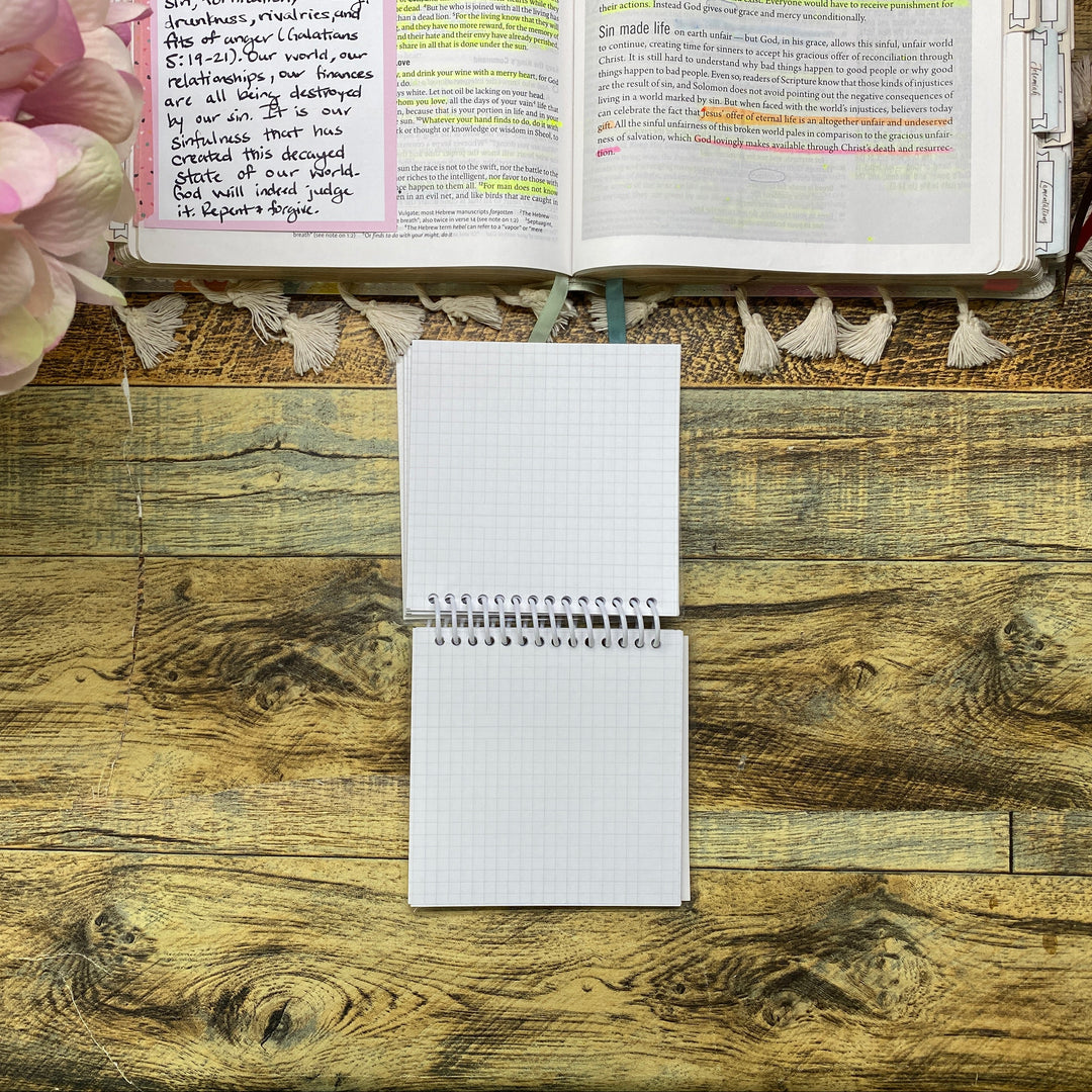 An open spiral-bound grid notebook lies on a rustic wooden tabletop next to an open book with handwritten notes and pink flowers in the corner.