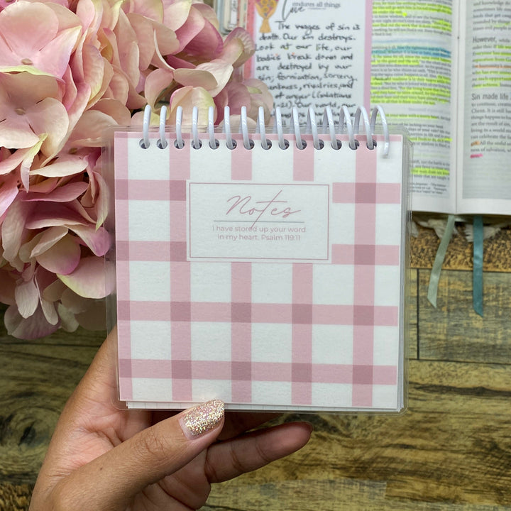 A hand holds a small pink and white gingham spiral notebook labeled 'Notes' above an open book with colorful highlighted pages, set on a wooden tabletop with pink flowers in the background.