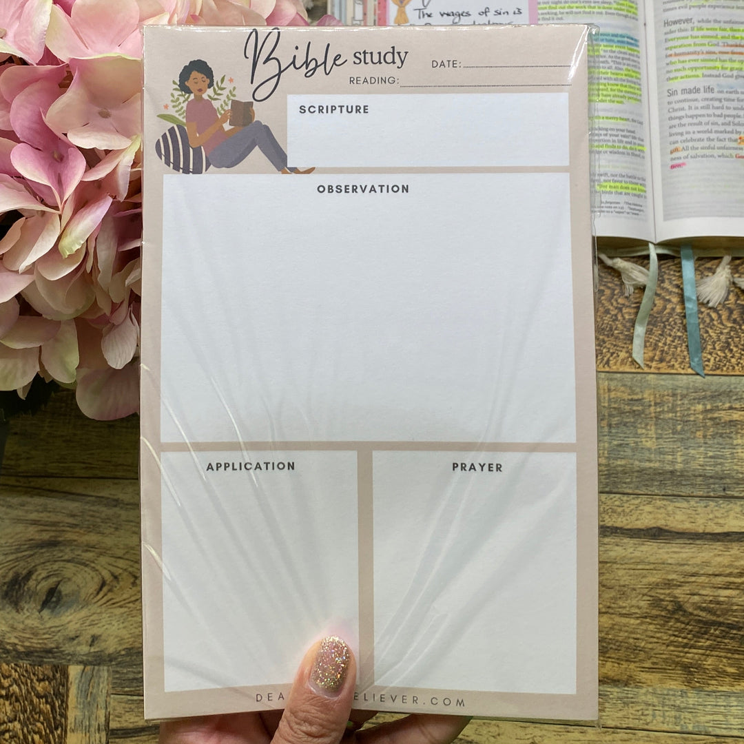 A hand holds a set of four white memo pads with a beige border and a small illustration on the top pad, placed on a wooden desk next to an open book and pink flowers.