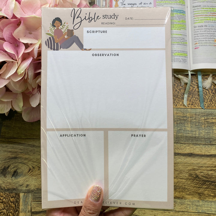 A hand holds a set of four white memo pads with a beige border and a small illustration on the top pad, placed on a wooden desk next to an open book and pink flowers.