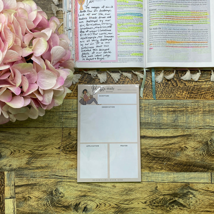 An open planner or journal with handwritten notes and colored highlight lines sits on a rustic wooden surface next to a blank page insert. A large pink hydrangea flower arrangement is visible at the top, with part of another book or notebook at the upper left.
