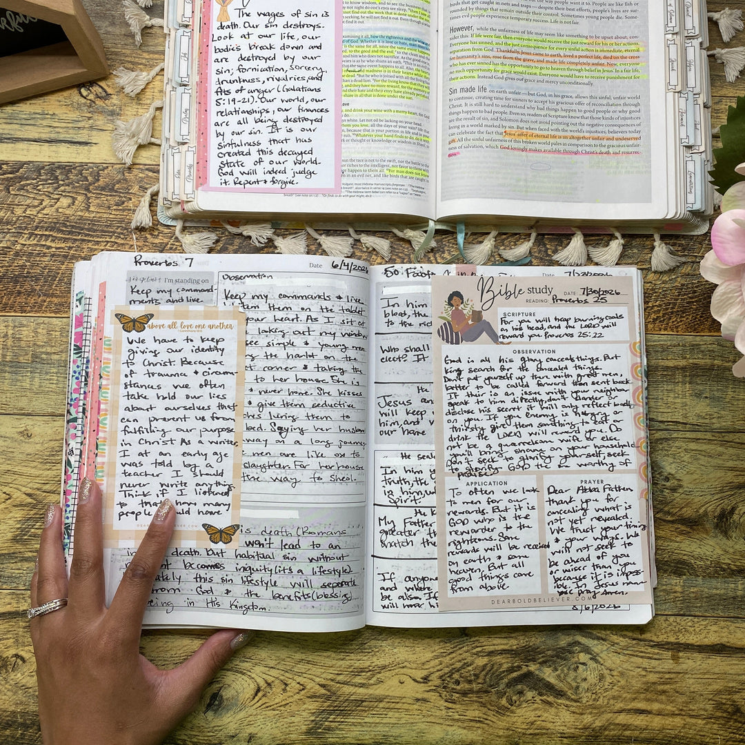 Two open handmade booklets or notebooks with handwritten pages and colorful highlights, displayed on a rustic wooden table. A hand is pointing at the left booklet, and pink flowers are visible in the foreground.