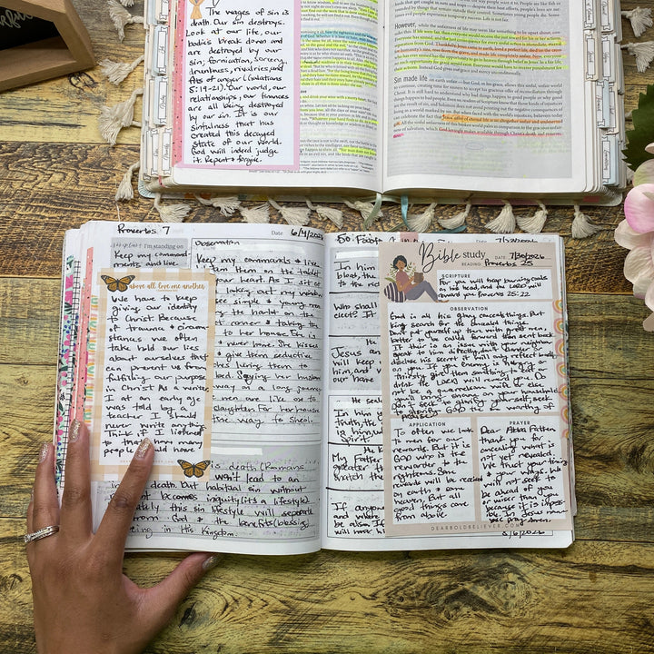Two open handmade booklets or notebooks with handwritten pages and colorful highlights, displayed on a rustic wooden table. A hand is pointing at the left booklet, and pink flowers are visible in the foreground.
