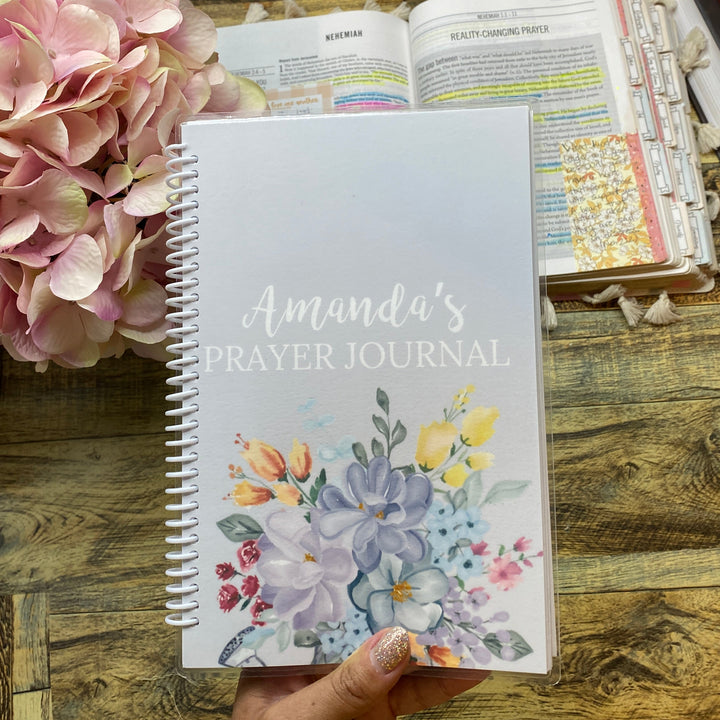 A hand holds a spiral-bound prayer journal titled 'Amanda's Prayer Journal' with a floral cover design, set on a rustic wooden table next to pink hydrangeas and other stationery items.