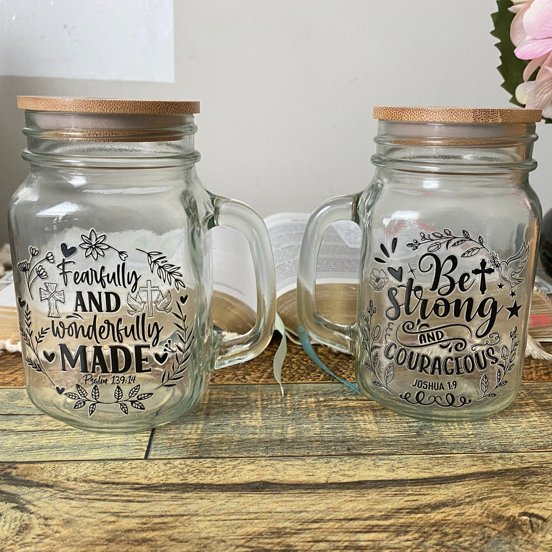 Two clear glass mason jar mugs with bamboo lids, displayed on a rustic wooden table. The left mug features the hand-lettered black print 'Fearfully AND wonderfully MADE', and the right mug features 'Bet Strong AND COURAGIOUS'.