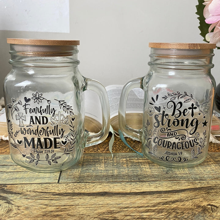 Two clear glass mason jar mugs with bamboo lids, displayed on a rustic wooden table. The left mug features the hand-lettered black print 'Fearfully AND wonderfully MADE', and the right mug features 'Bet Strong AND COURAGIOUS'.