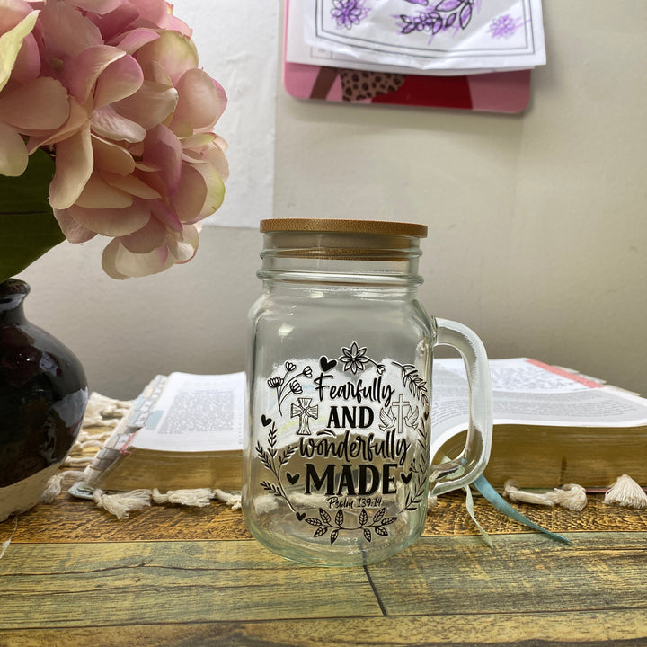 A clear glass mason jar with a wooden lid and handle, printed with the phrase 'Frequently and wonderfully MADE' in black lettering, set on a rustic wooden table next to an open book and a vase of pink flowers.