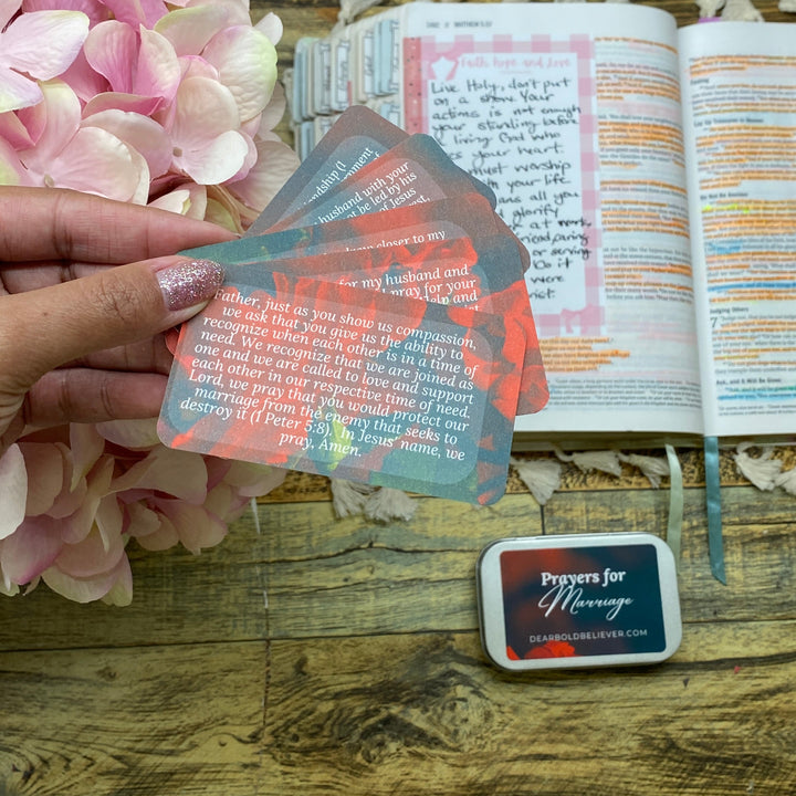 A hand holding a translucent pink and blue stamp ink pad over an open journal, with a small square ink pad block nearby and pink flowers on a wooden tabletop.