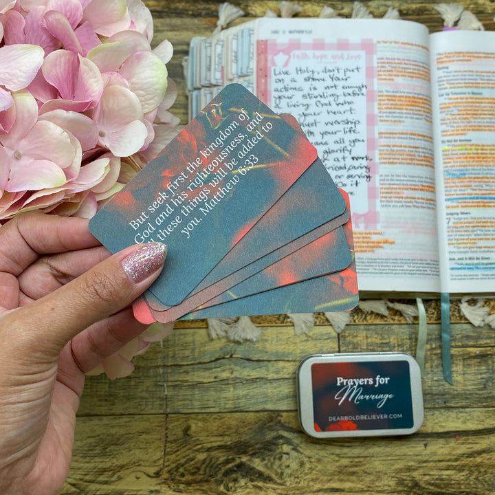 A hand holds a fan of printed cards over an open journal on a wooden table, with pink hydrangea flowers and a small ink pad visible in the scene.