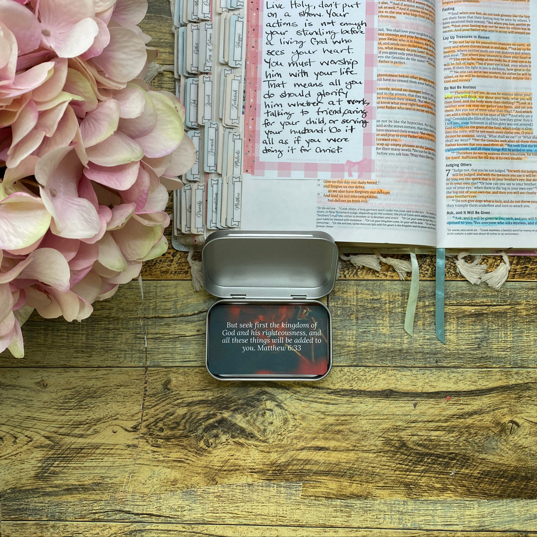 An open journal or notebook spread on a wooden surface, with handwritten notes, colorful printed inserts, a small metal tin, and a cluster of pale pink hydrangea flowers at the top of the frame.