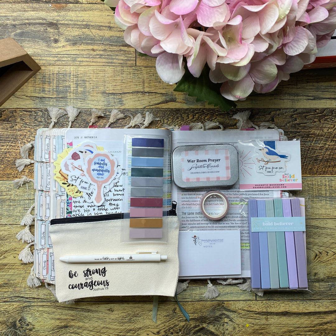 An open journal or planner spread on a rustic wooden table, showing a cream fabric pouch with a pen, a color palette strip, sticky notes, a small metal tin, pastel sticky notes, and other stationery items. A pink hydrangea and part of a book are visible at the right edge.