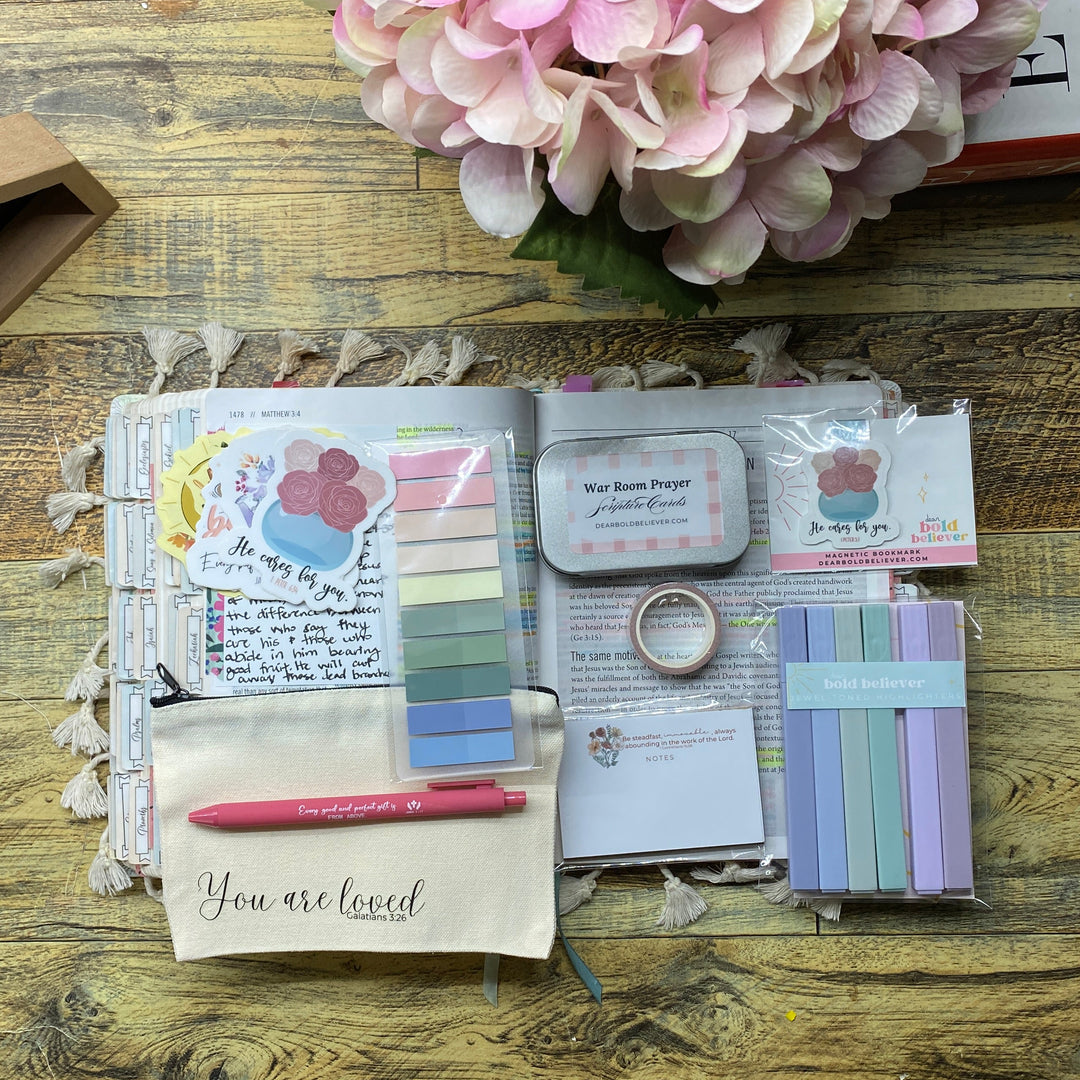 A flat-lay display of a stationery bundle containing a planner, sticky notes, washi tapes, a pink pen, and small cards on a rustic wooden table next to a pink hydrangea.