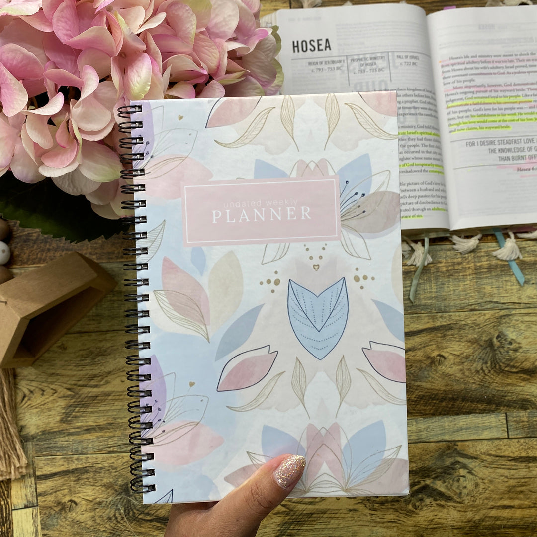 A hand holds a spiral-bound planner with a pastel floral cover on a rustic wooden table, next to an open book and pink flowers.